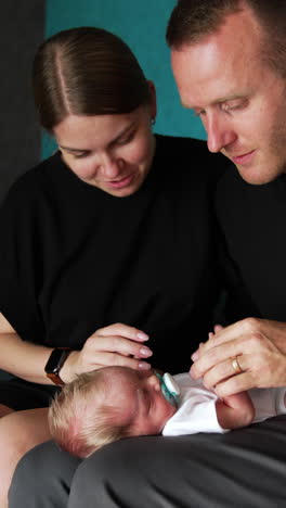 new-made parents admiring their cute newborn baby. kid lies on dad's laps sleeping and parents touch his tiny hands and nose. vertical video