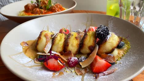 tasty syrniki with forest berries, banana and coconut shavings in cashew cream and honey, mini breakfast cheese pancakes and meatballs in the background, traditional ukrainian russian dishes, 4k shot