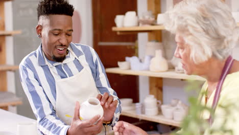 Two-happy-diverse-male-and-female-potters-holding-pots-and-discussing-in-pottery-studio,-slow-motion