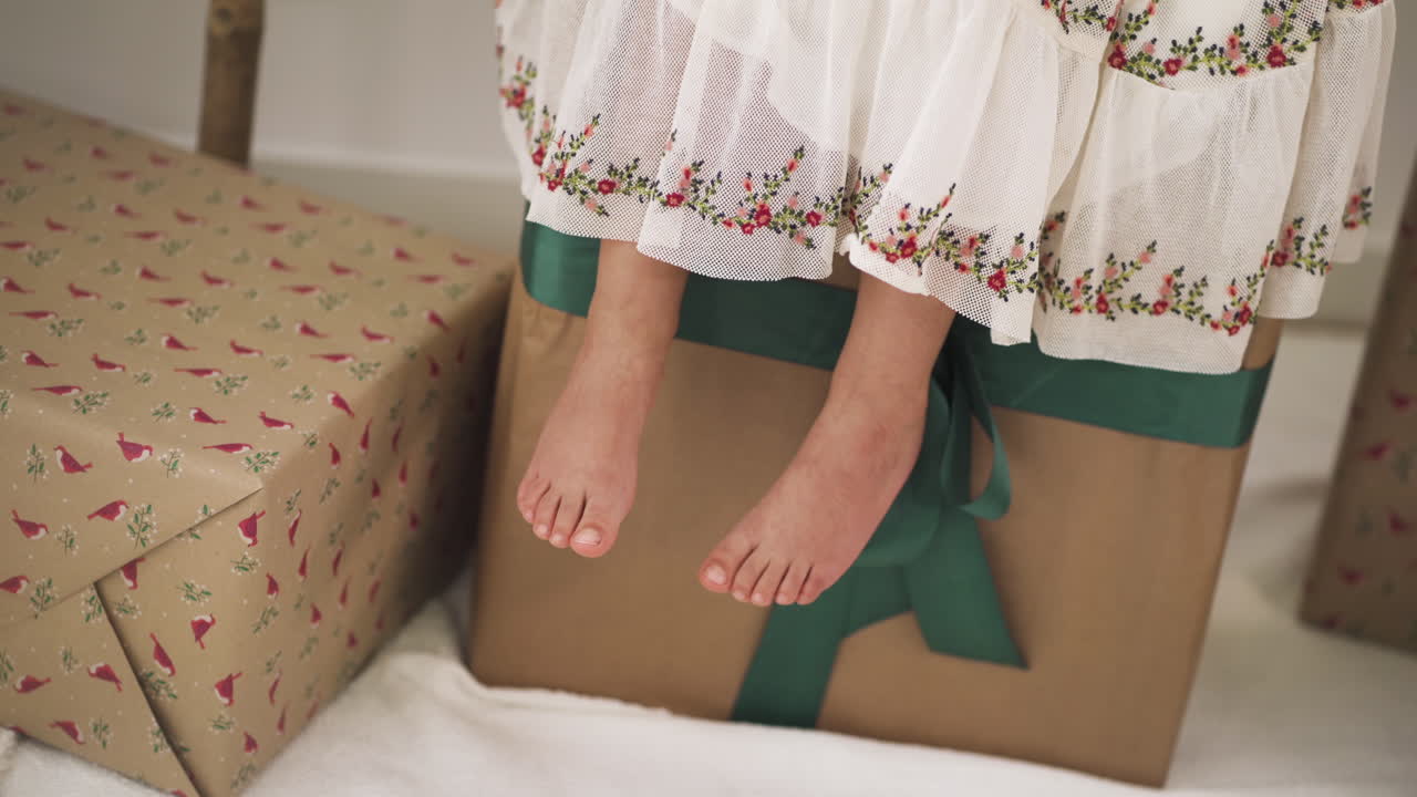 chica balanceando sus pies vestida con un vestido festivo sentada en una gran caja de regalo de navidad - primer plano de ángulo alto