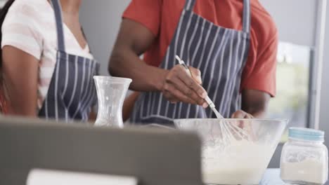 Happy-african-american-couple-in-aprons-baking-together-using-tablet-in-kitchen,-slow-motion