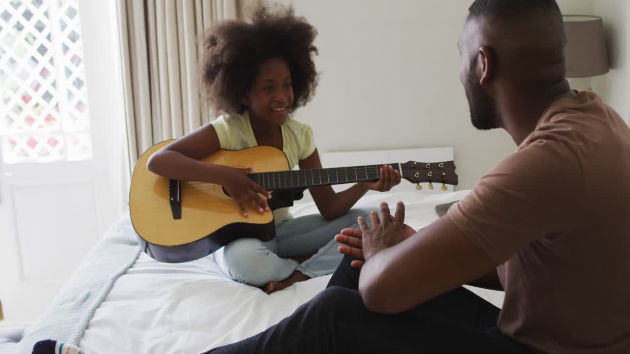 African american daughter playing guitar sitting on bed with her father listening and clapping
