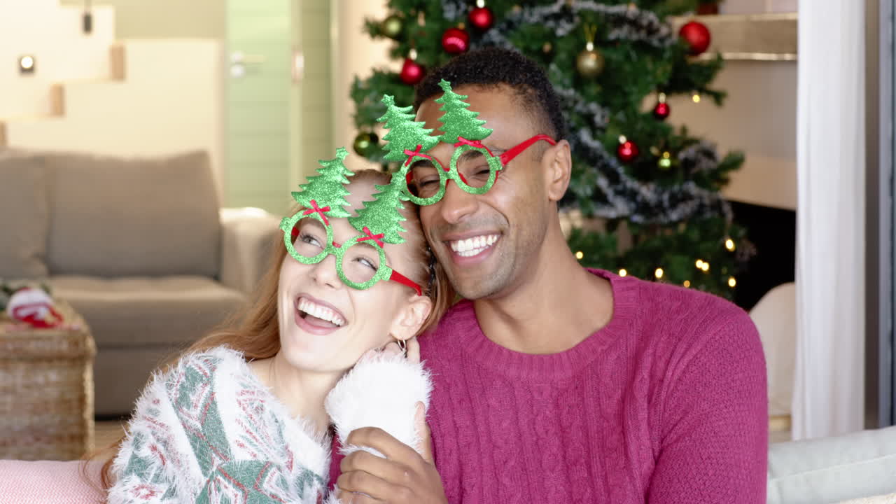 Pareja sonriente con gafas festivas disfrutando la Navidad en casa junto al árbol.