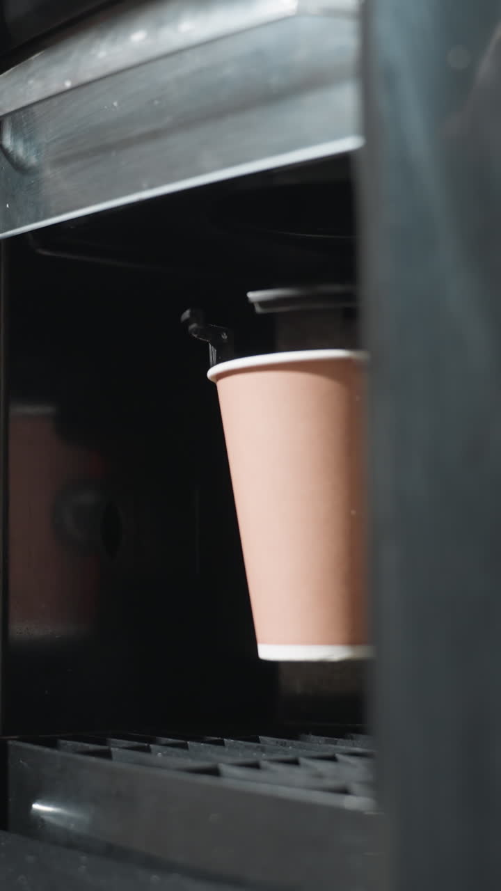 Vaso de papel recibiendo café caliente dentro del dispensador de una máquina expendedora con zona de caída del vaso visible, capturando la bebida caliente que se sirve en un entorno de oficina moderno con una planta borrosa al fondo