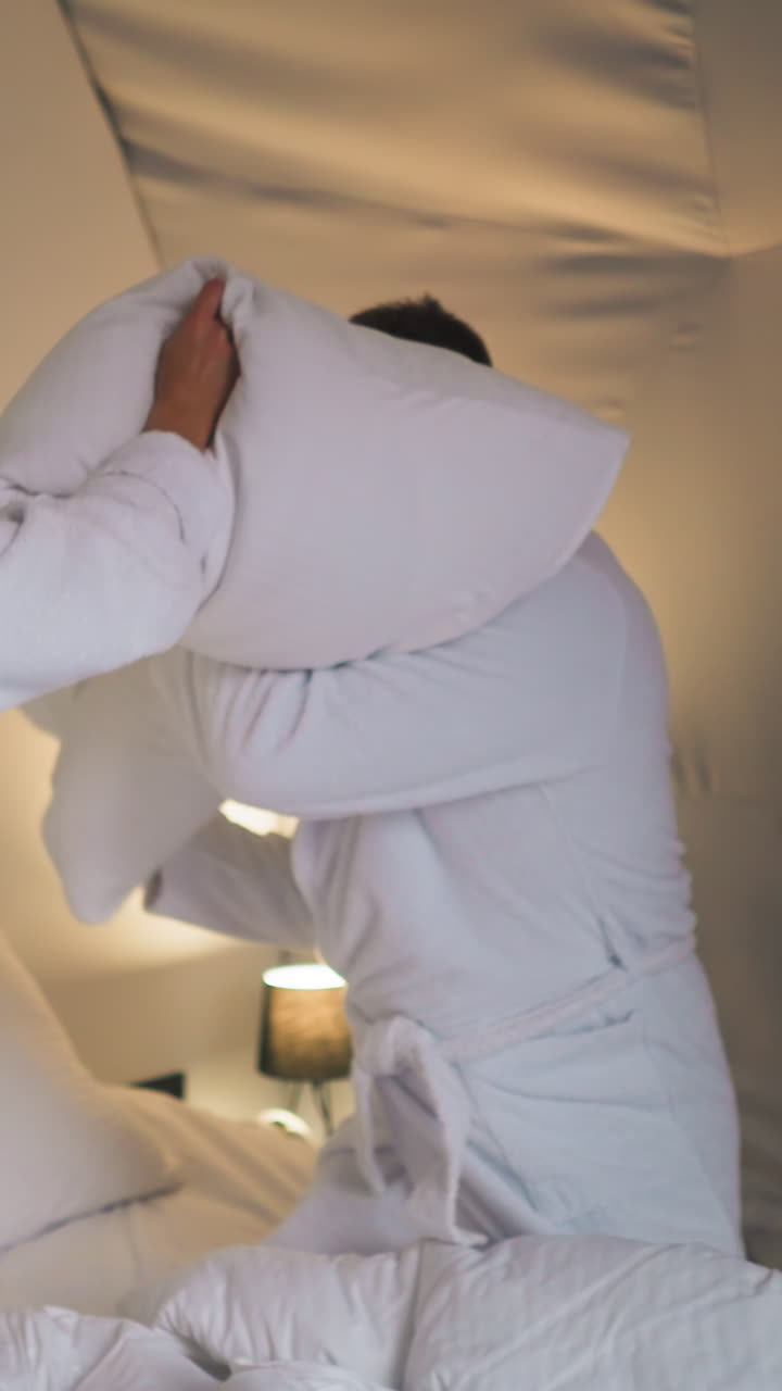 Newlyweds engage in playful pillow fight on bed. Young people in soft bathrobes enjoy funny battle in glamping house. Memorable honeymoon trip
