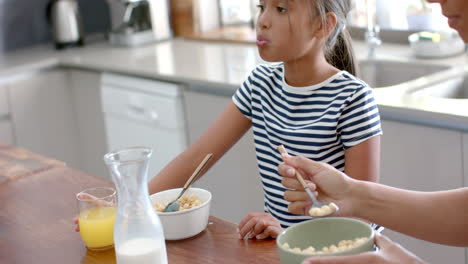 madre y hija biraciales comiendo cereales para el desayuno en una cocina soleada, cámara lenta
