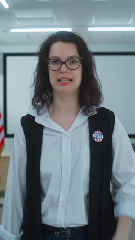 American-female-voter-or-polling-worker-with-badge-walks-and-speaks-on-camera,-calls-for-voting.-National-Election-Day-in-the-United-States.-Voting-booths-at-polling-station.-Civic-duty-and-patriotism