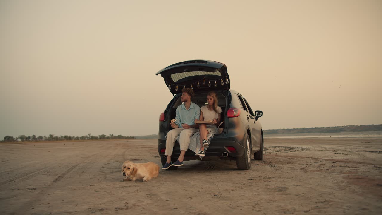un hombre barbudo con cabello rizado en una camisa azul y su novia rubia están comiendo pizza y sentados en el maletero del coche negro decorado con las luces contra el cielo amarillo, mientras su perro está acostado y comiendo en el suelo