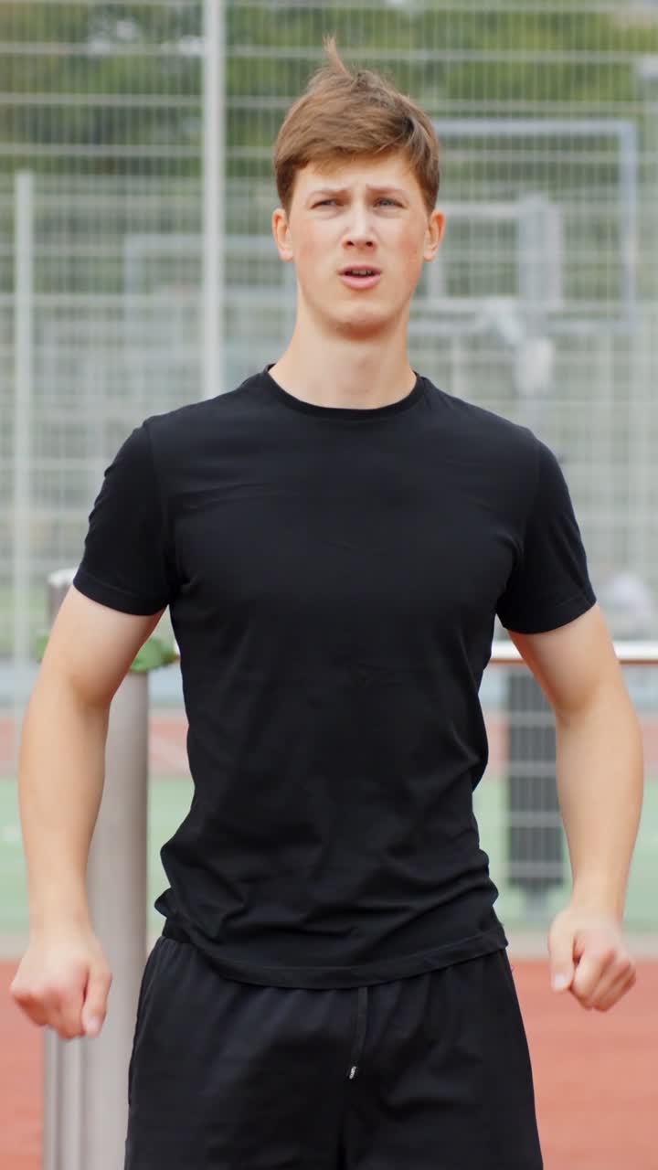 Young man in athletic black wear stretching arm across chest, performing a warm up exercise on an outdoor sports track, focusing on physical preparation for his workout, vertical static slow motion