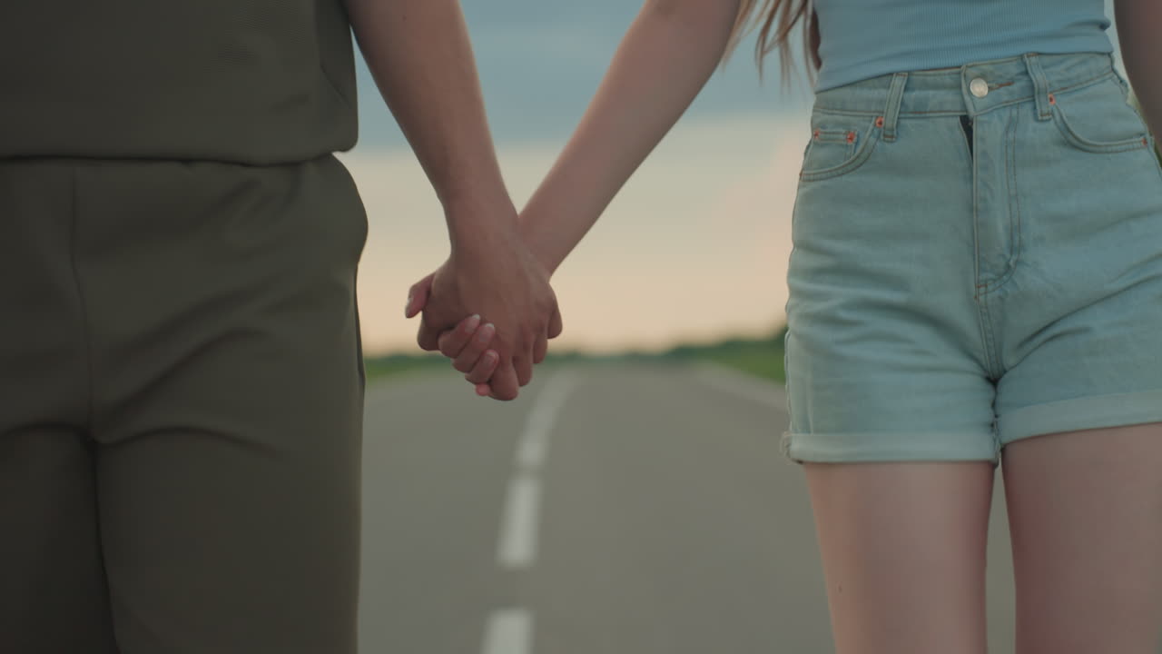 close up rear view of couple holding hands walking down empty rural road under dramatic sunset sky with soft clouds green fields on horizon conveying romance adventure partnership