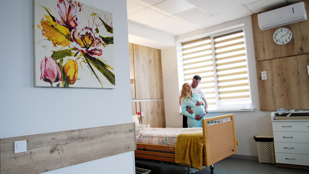 Cozy comfortable ward for couples expecting baby. Couple stands at the bed embracing and smiling.