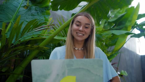 a girl in a blue dress remotely online working behind a laptop and looking into the screen the backyard with green plants