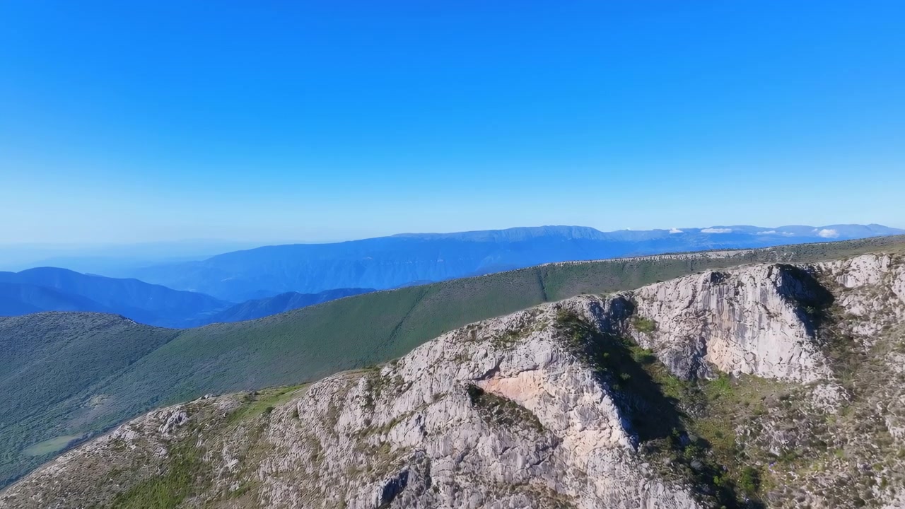 Vista aerea obliqua elevata di una cresta calcarea frastagliata con ripide scogliere erose e rada vegetazione arbustiva in primo piano, che conduce a dolci pendii erbosi e colline ondulate che si trasformano in
