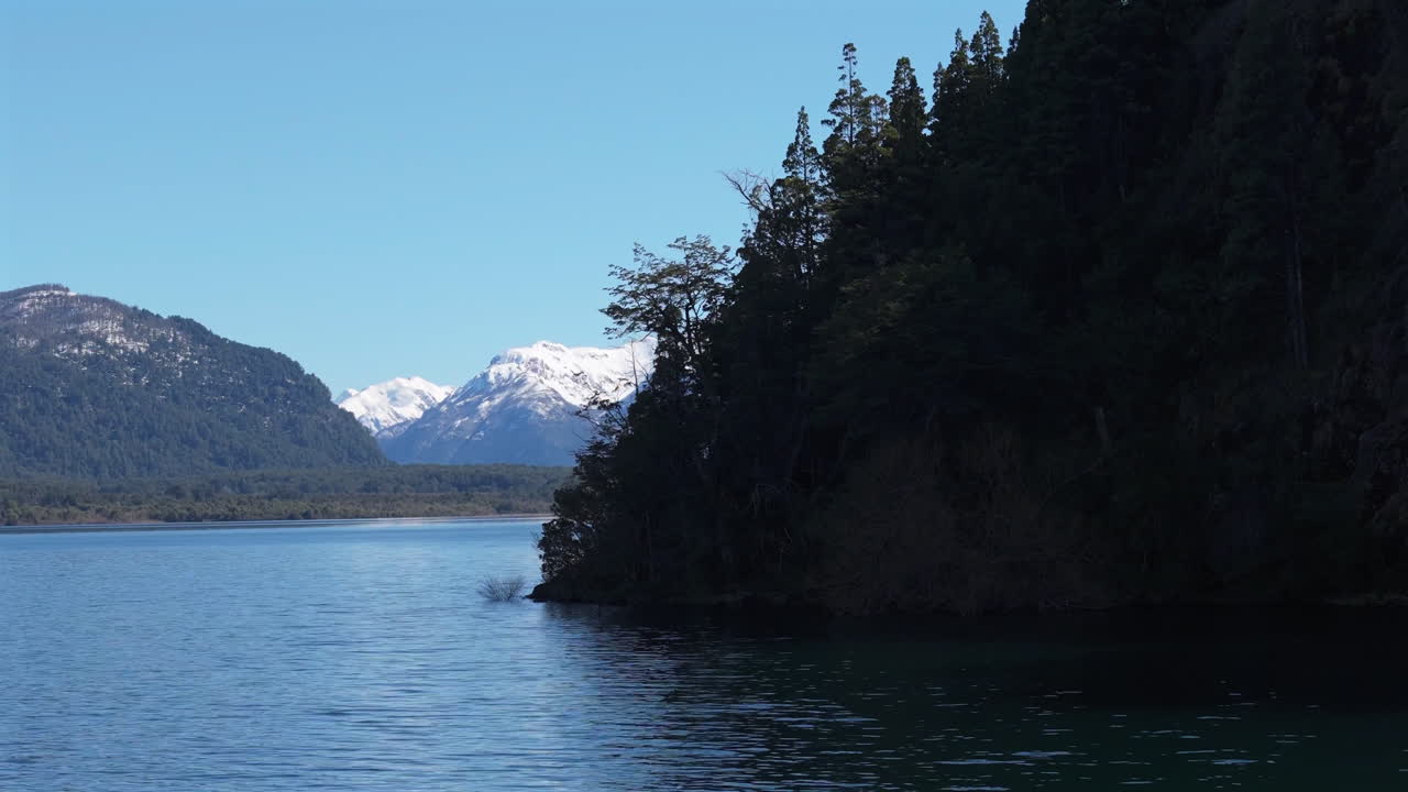 Forward tracking drone shot over waters in Lago Futalaufquen, Parque Nacional los Alerces. Snowed peaks in back contrast with blue sky and dark trees. Shot on 4K-60fps.
