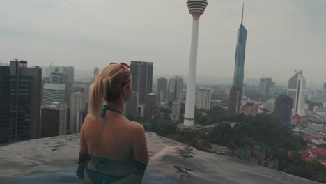 a blonde girl in an infinity pool looks out over the city of kuala lumpur