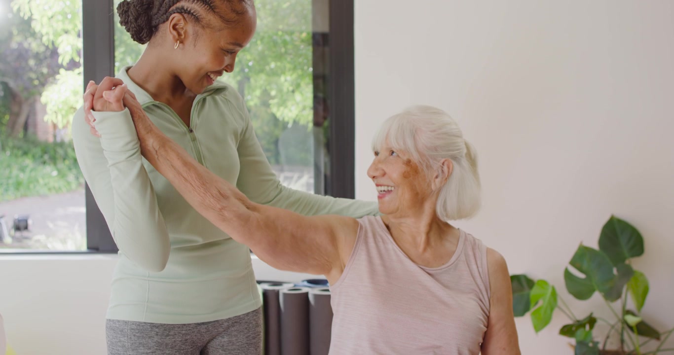 Diverse caregiver and senior woman responding to cue, guiding arm stretch on chair in living room
