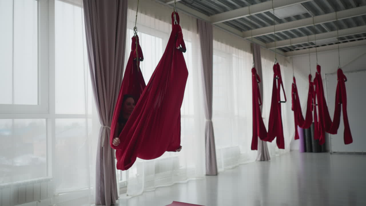 Bright yoga studio with glass windows as yoga specialist wraps body within red aerial silk hammock swinging gently, demonstrating flexibility strength focus serene practice above mat