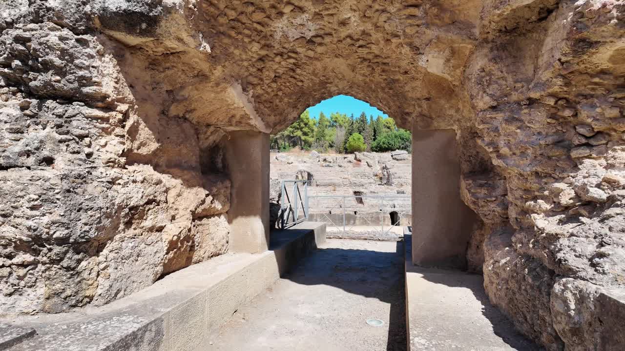 Vista attraverso un arco di pietra grezza che rivela le rovine dell'antico anfiteatro romano nel sito archeologico di Italica vicino a Siviglia, Spagna. Strutture in pietra storiche e architettura classica.