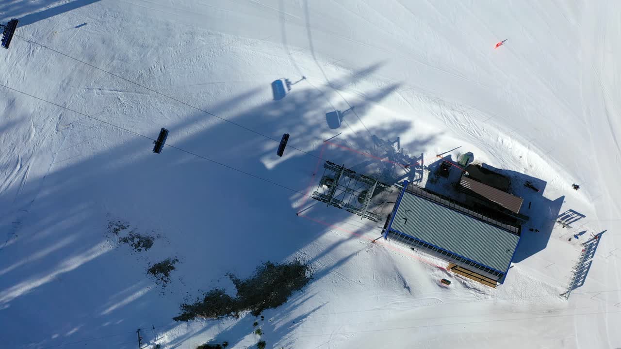 High drone shot of a chair lift moving above skiers and fresh powder, cutting through pine forests on a winter mountain, with majestic peaks in view under a crisp, clear sky
