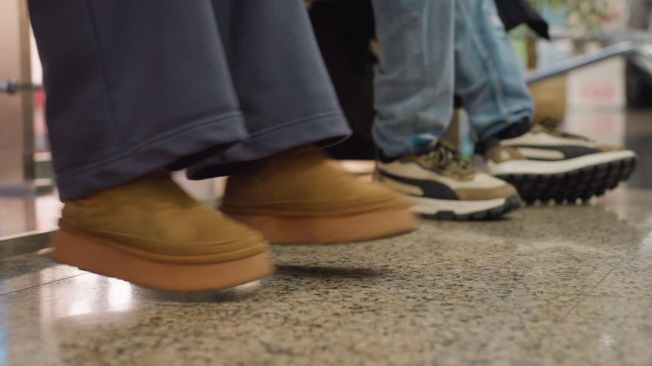 Side view of seated individuals on polished granite floor with focus on legs, one person swinging feet in brown suede shoes while others sit still, showing casual style