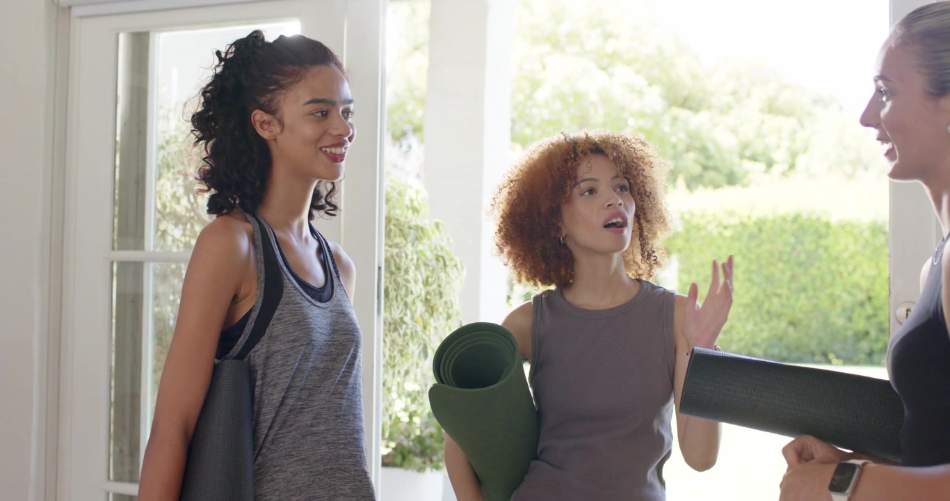 Diverse female friends and host arriving home entry, speaking and gesturing holding mats for class