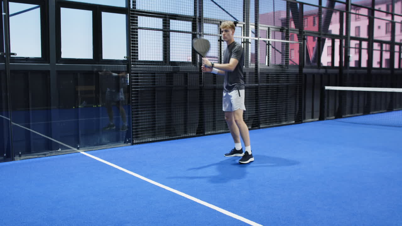 Young man playing padel tennis, preparing to hit ball on blue indoor court