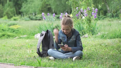 a nine-year-old girl after school sits in a park on the grass and plays a game on her smartphone. she shows joyful hand gestures and reacts emotionally.