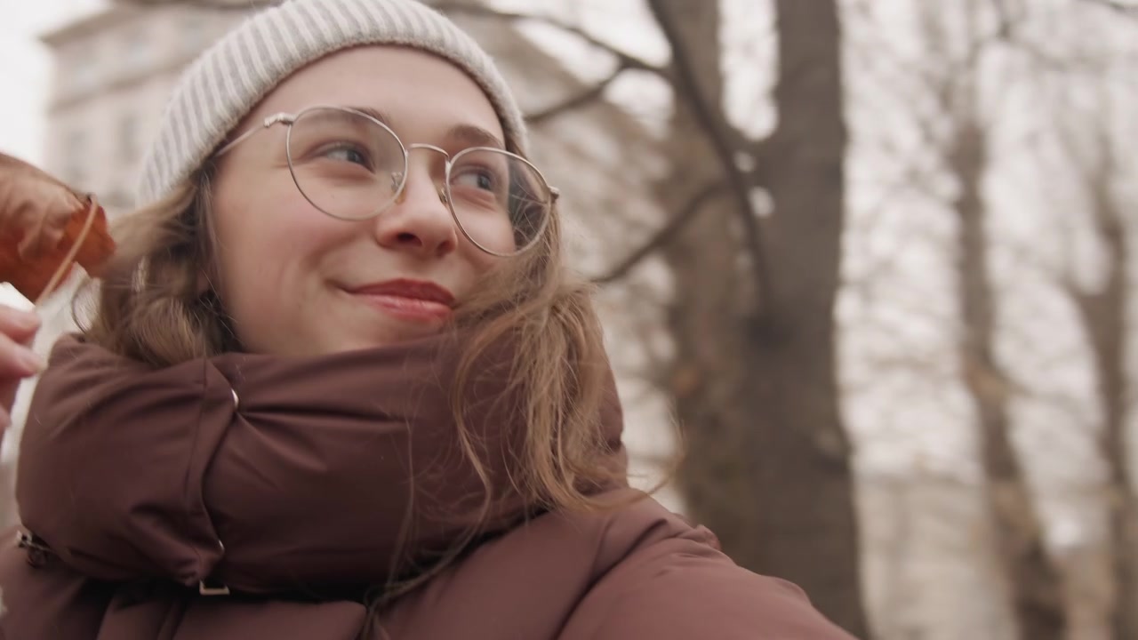 Donna bianca content creator sorridente, foglia in primo piano, atmosfera da selfie dal basso con occhiali e berretto di lana, espressione calda e gesto giocoso, piumino e alberi spogli che incorniciano lo sfondo urbano, accogliente