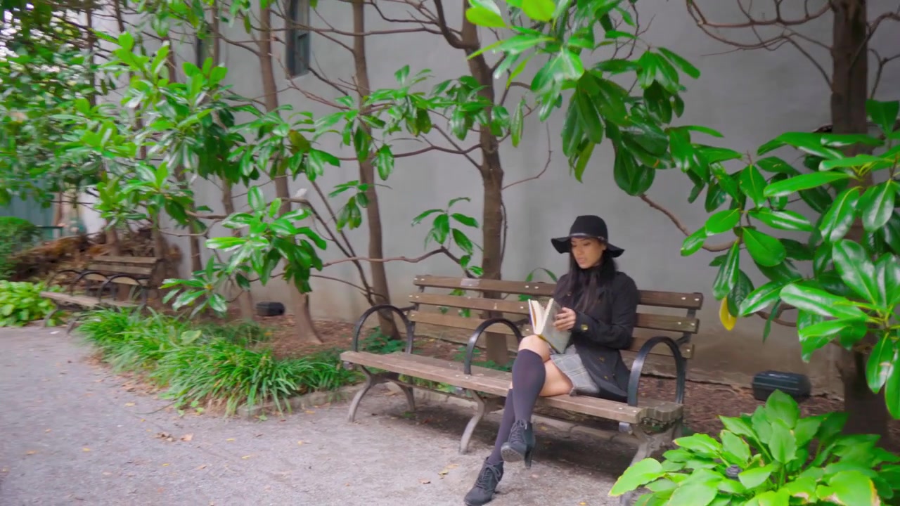 Seorang wanita muda, tenggelam dalam pikiran, berjalan melalui taman yang rimbun dan hijau. Sebuah buku dipegang lembut di tangannya saat dia membenamkan diri dalam lingkungan alam yang tenang