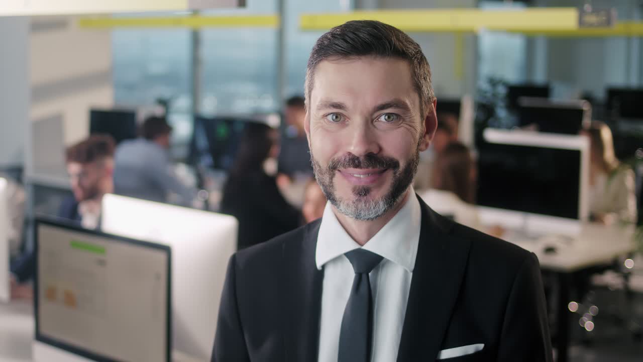 Portrait of adult business man boss standing in office smiling looking at camera while employees are busy working in background