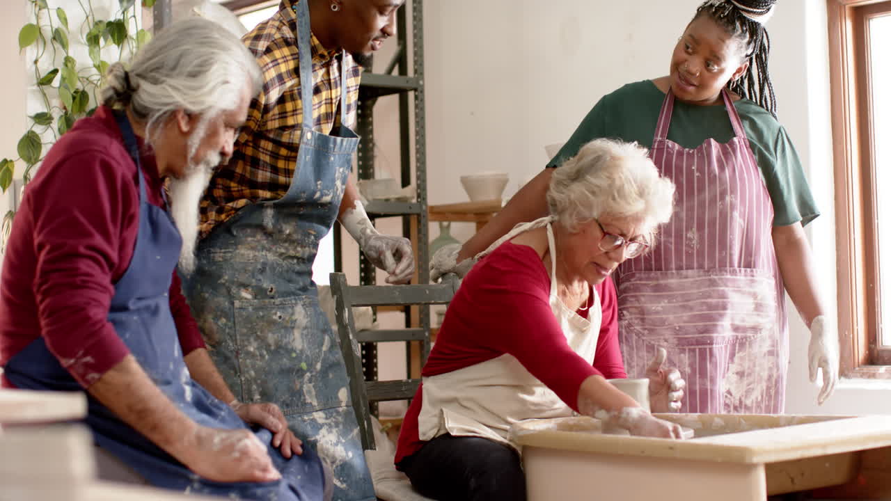 Happy biracial female potter with others, using potter's wheel in pottery studio, slow motion