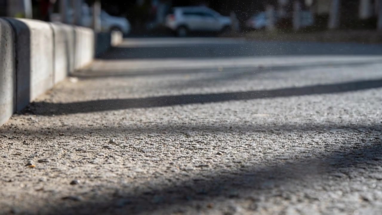 La telecamera panoramica si abbassa rivelando asfalto ruvido con lunghe ombre, un marciapiede e un SUV bianco per i dettagli della strada.