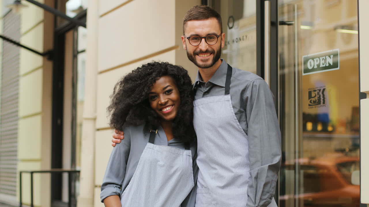 retrato de camareros femeninos y masculinos abrazándose y sonriendo a la cámara fuera de una cafetería moderna