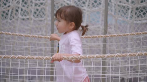 toddler exploring rope bridge in indoor playground, having fun and staying active as she hops and bounces grabbing onto each side, tracking sideview slow motion