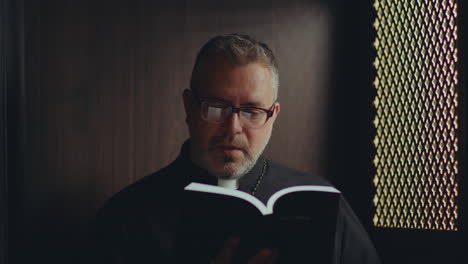 catholic priest reading bible and praying in confessional booth