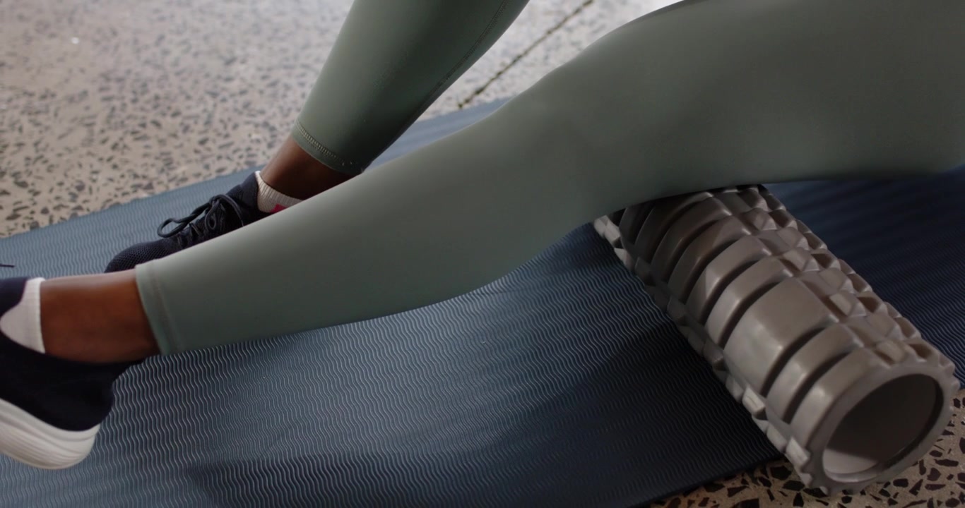 African American 30s woman targeting ache placing foam roller under right thigh on blue mat rolling
