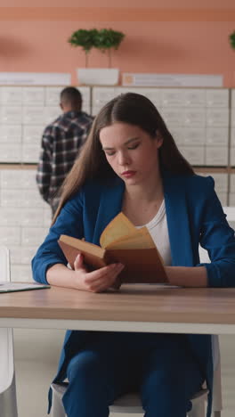 woman reads book in library hall. thoughtful student learns educative material sitting at table in college reading room. literature usage for studying