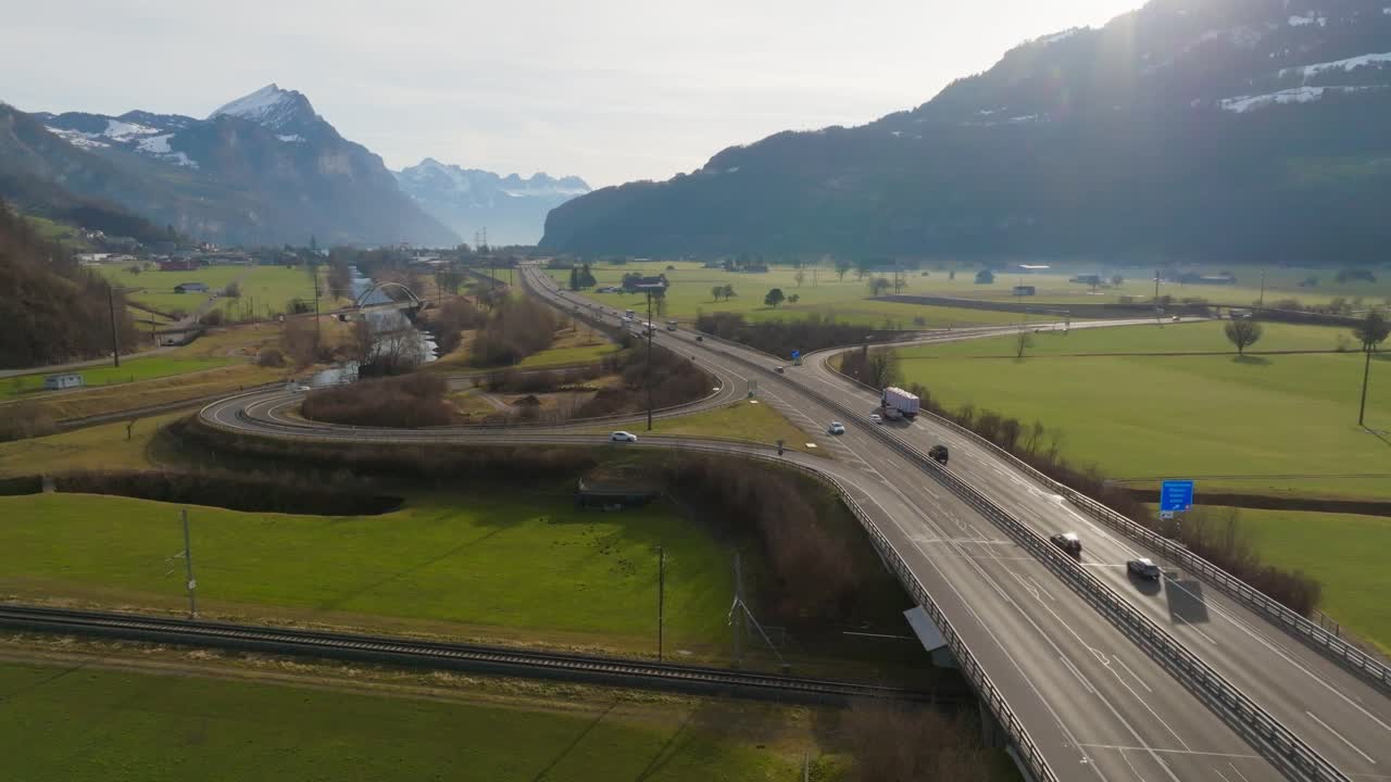Drone flight parallel to a highway running through a green mountain valley. Cars and trucks move along the road under clear daylight, surrounded by fields and distant alpine peaks