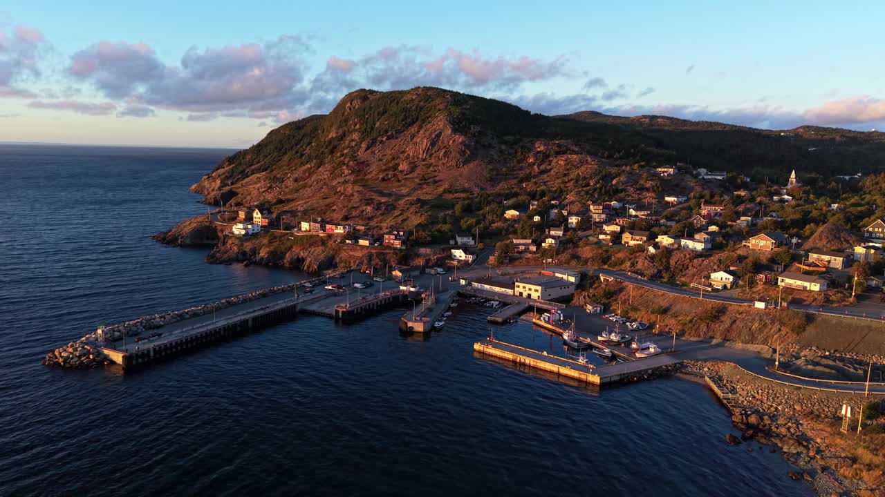 Golden-hour aerial shot of Portugal Cove’s harbor and rugged hillside in Newfoundland, capturing ocean waves against rocky shoreline and the village bathed in warm light