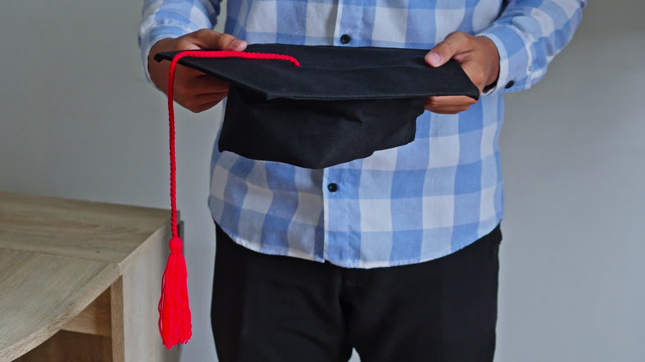 Young man holding square academic cap near wooden desk