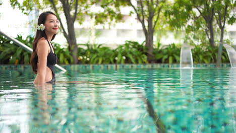 Beautiful-young-Asian-woman-with-white-flower-in-hair-seated-in-warm-clear-swimming-pool-with-waterfall-in-background