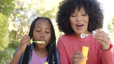 Happy-african-american-mother-with-daughter-blowing-bubbles-in-garden-at-home,-slow-motion