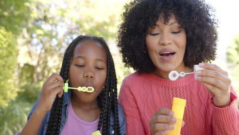 feliz madre afroamericana con su hija soplando burbujas en el jardín en casa, cámara lenta