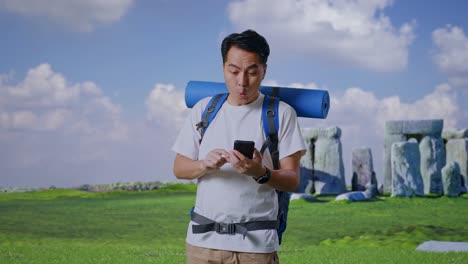 asian male hiker with mountaineering backpack looking at the smartphone and saying wow while traveling in stonehenge