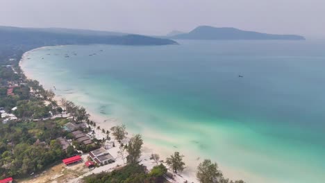 aerial view of kaoh rong sanloem, cambodia featuring white sandy beaches, lush greenery, clear turquoise waters, and scattered boats along the calm coastline with distant hills fading into the horizon
