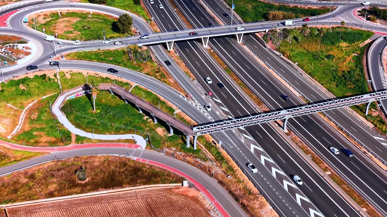Aerial view of a highway interchange with pedestrian bridge, Spain. Multiple traffic lanes, roundabouts and a curved walkway wind through the landscape.