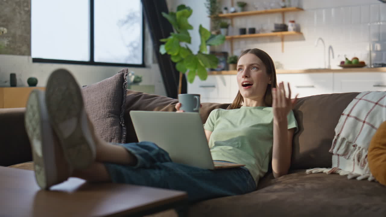 Home girl videocalling laptop lying comfortable apartment sofa with coffee cup