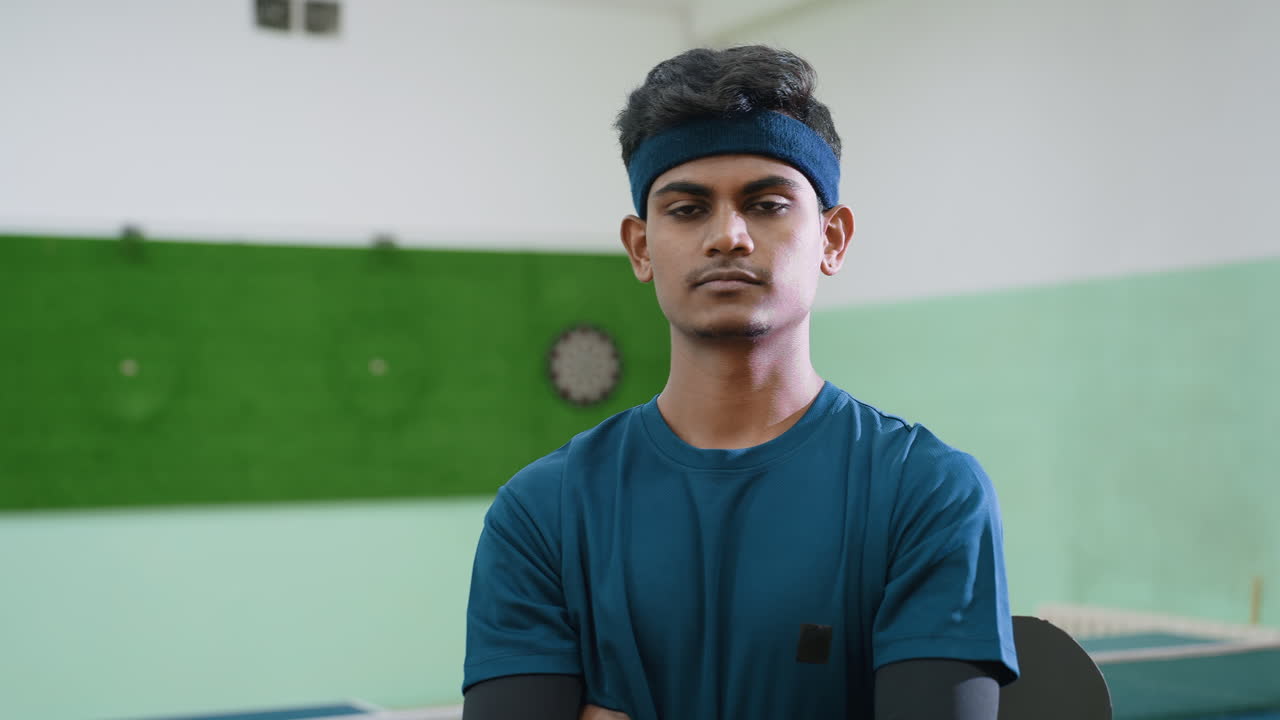 Confident tennis trainer in blue sportswear stands indoors holding racket, folding arms with serious expression, showing focus, strength, and readiness for professional training session on court
