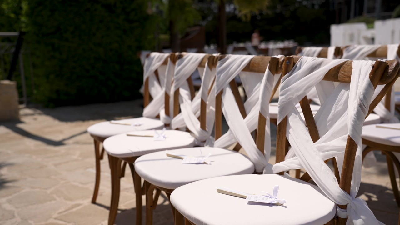 Filas de sillas rústicas de madera están cuidadosamente dispuestas para una ceremonia de boda. Cada silla está adornada con tela blanca y delicados detalles, creando un ambiente acogedor para los invitados