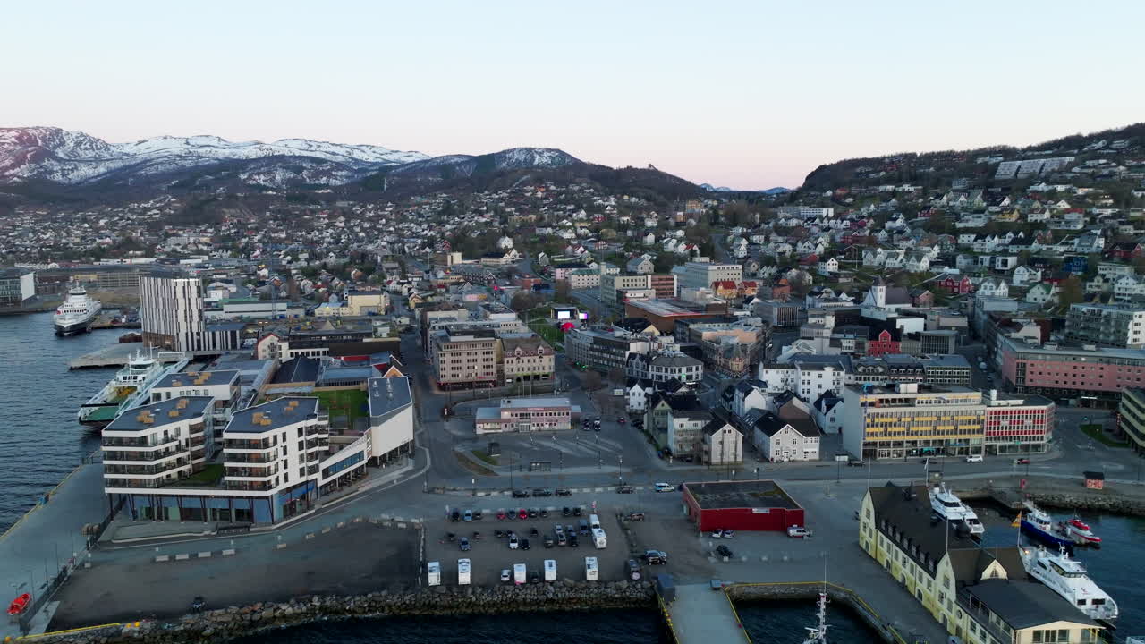 Harstad city waterfront and harbor at sunset, modern architecture, boats, and snow-capped mountains in northern Norway. Aerial forward